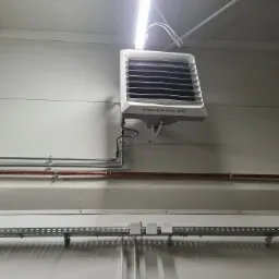 Zamontowany nagrzewnica wodna Volcano EC z widocznymi rurami instalacji grzewczej, umieszczona na białej ścianie w hali. Widoczna instalacja elektryczna i oświetlenie.