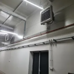 Dwa przemysłowe nagrzewnice powietrza VOLCANO EC zamontowane na ścianie hali, widoczne rury i kable instalacyjne, oświetlenie LED sufitowe.