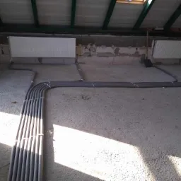 Szare rury instalacyjne ułożone na surowym betonowym podłożu w pomieszczeniu z dwoma grzejnikami na ścianach i ukośnym sufitem poddasza.