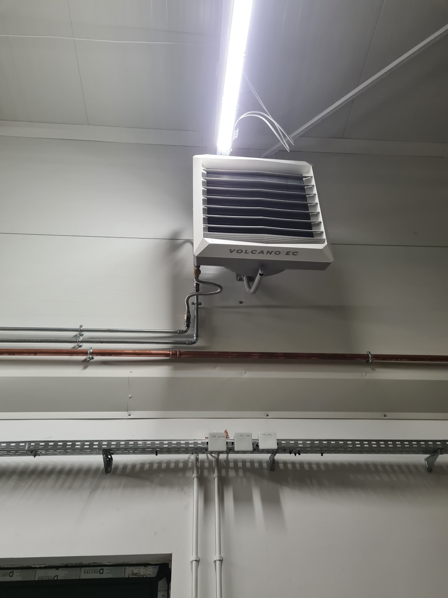 Zamontowany nagrzewnica wodna Volcano EC z widocznymi rurami instalacji grzewczej, umieszczona na białej ścianie w hali. Widoczna instalacja elektryczna i oświetlenie.