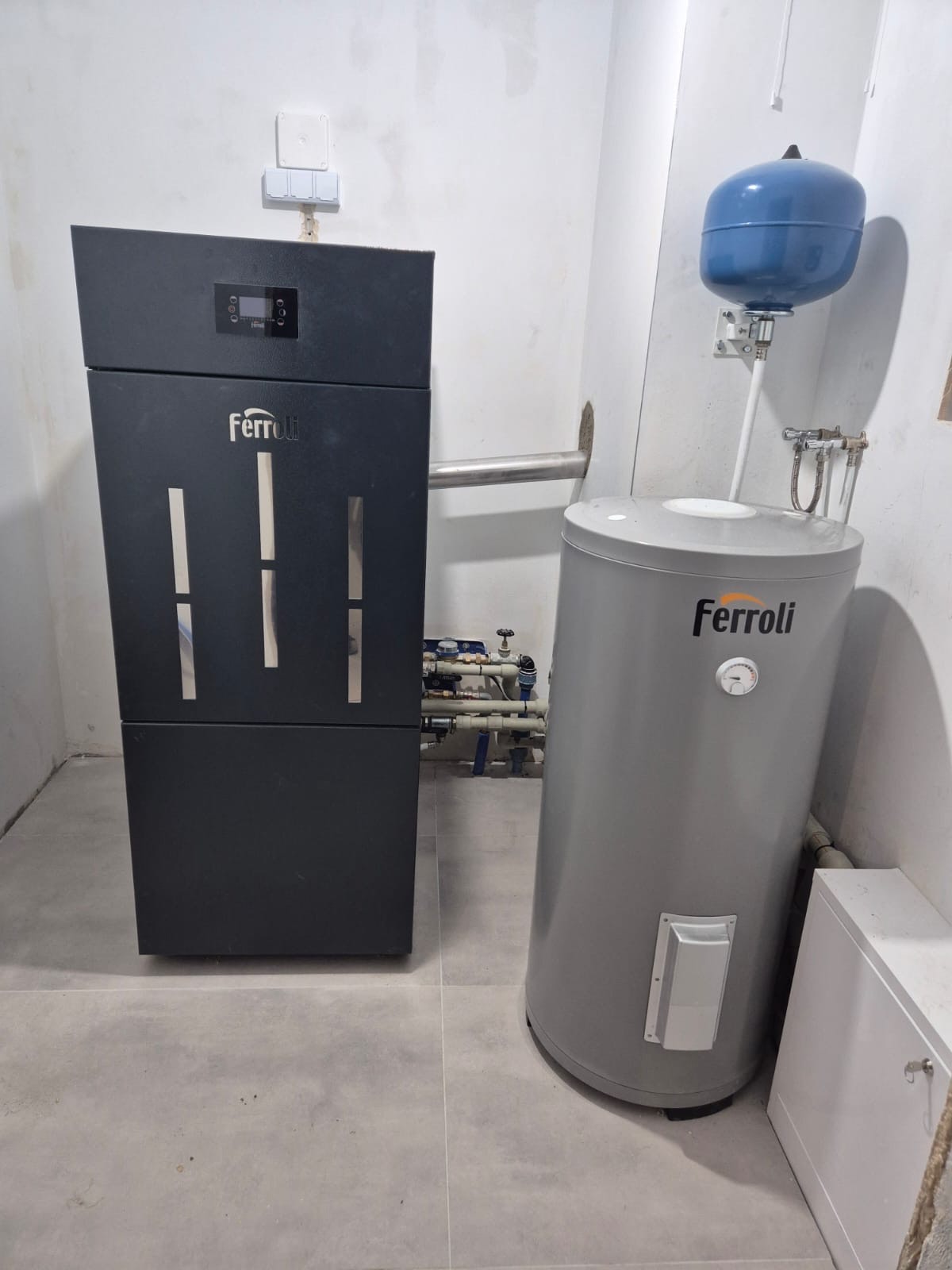 Czarny piec Ferroli obok szarego zasobnika wody z niebieskim naczyniem wzbiorczym. Widoczne rury i instalacja hydrauliczna. Szare płytki na podłodze.