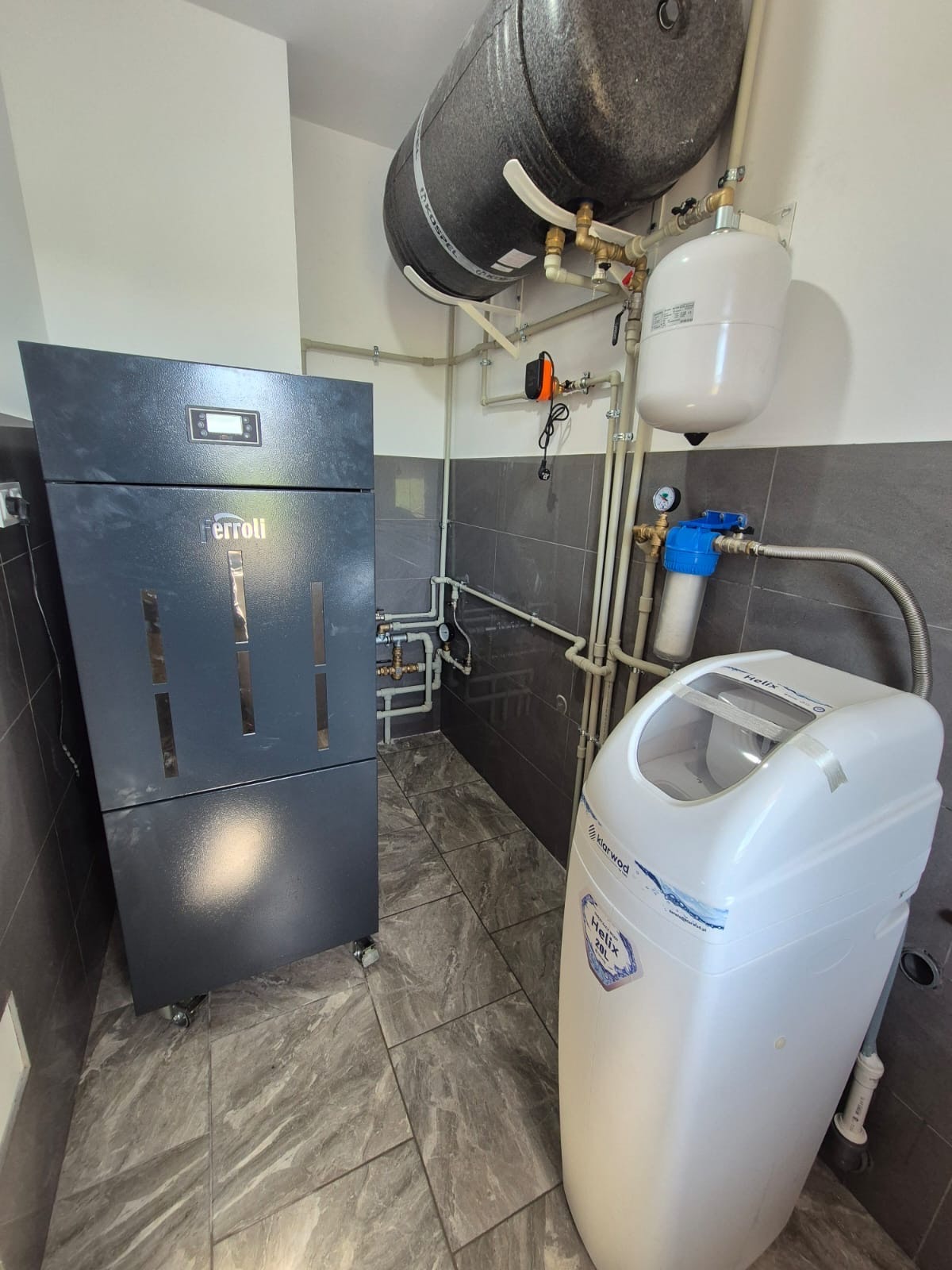 Zainstalowany piec Ferroli z podłączonym zbiornikiem i systemem uzdatniania wody w kotłowni z szarymi płytkami. Widoczne rury i zawory, instalacja grzewcza.
