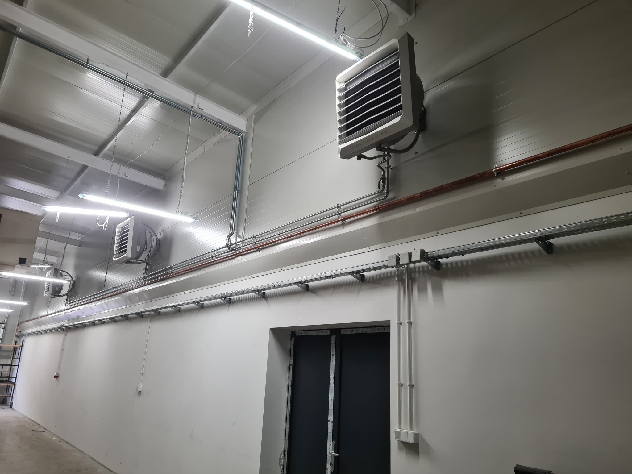 Dwa przemysłowe nagrzewnice powietrza VOLCANO EC zamontowane na ścianie hali, widoczne rury i kable instalacyjne, oświetlenie LED sufitowe.