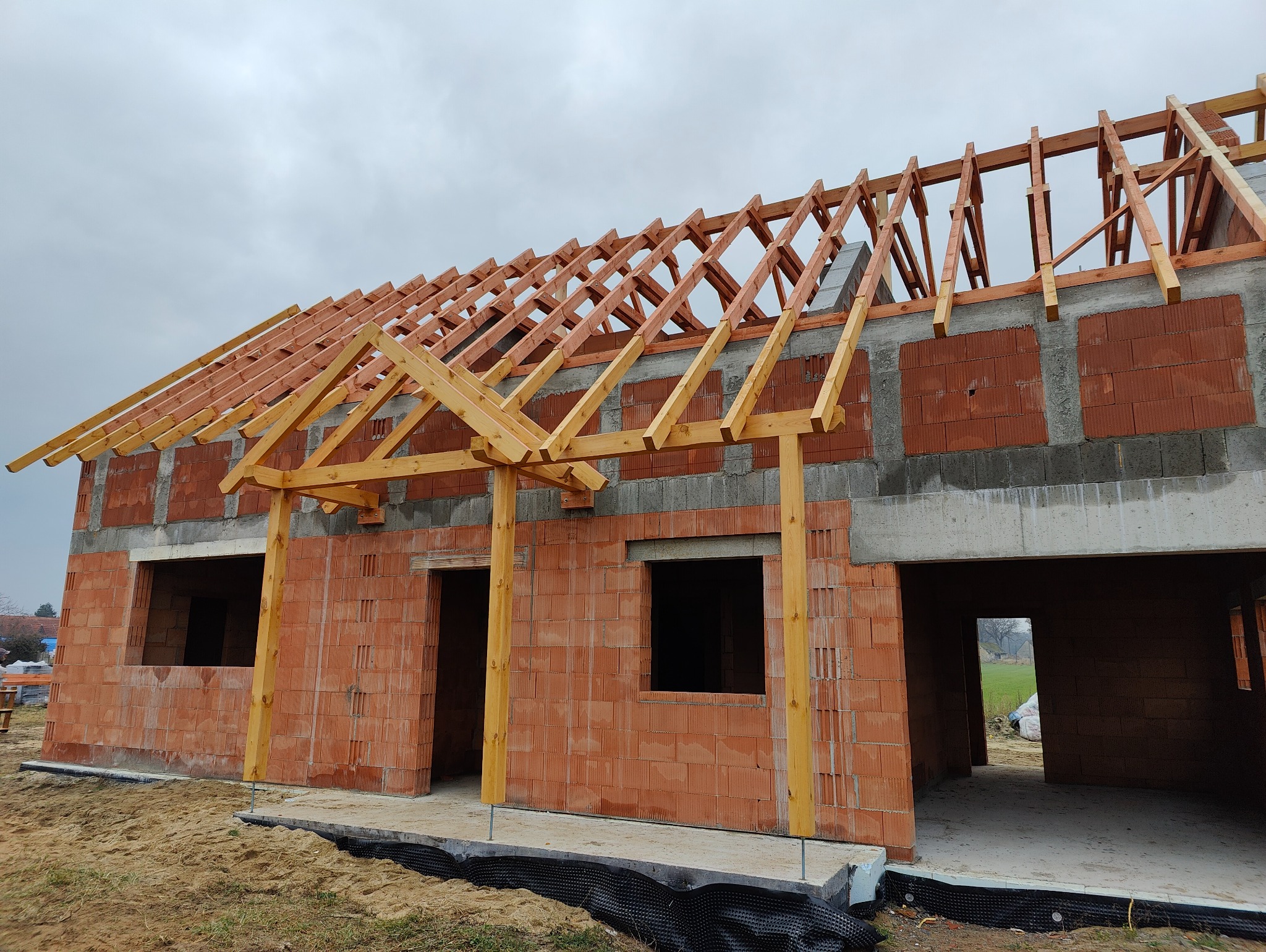 Surowy stan budynku z czerwonej cegły z widoczną drewnianą konstrukcją szkieletu dachu na pochmurnym tle.