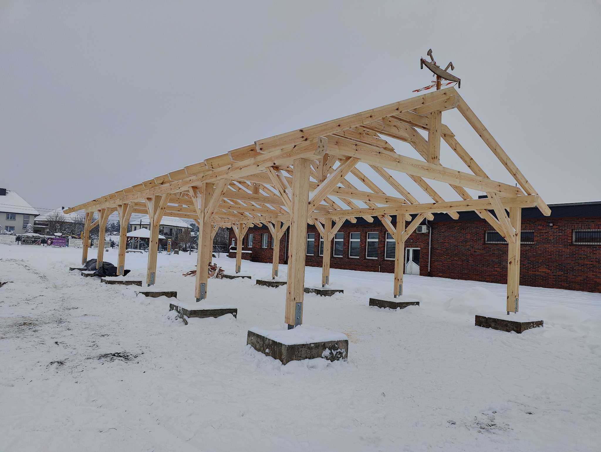 Surowa, drewniana konstrukcja z więźbą dachową w trakcie budowy, widoczna na tle zasypanego śniegiem terenu i ceglanego budynku, z dekoracyjnym wiatrowskazem na szczycie.