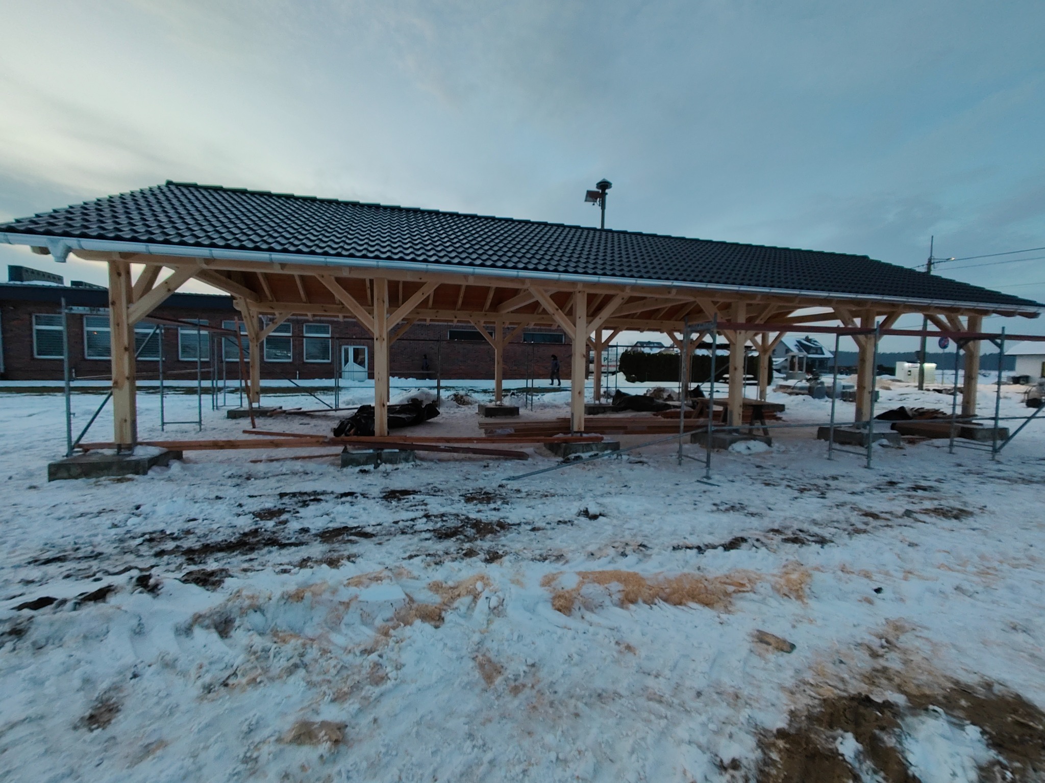 Budowa zadaszenia z drewnianą konstrukcją i czarnym pokryciem dachowym w zimowym krajobrazie, widoczne rusztowania i przygotowane elementy drewniane.