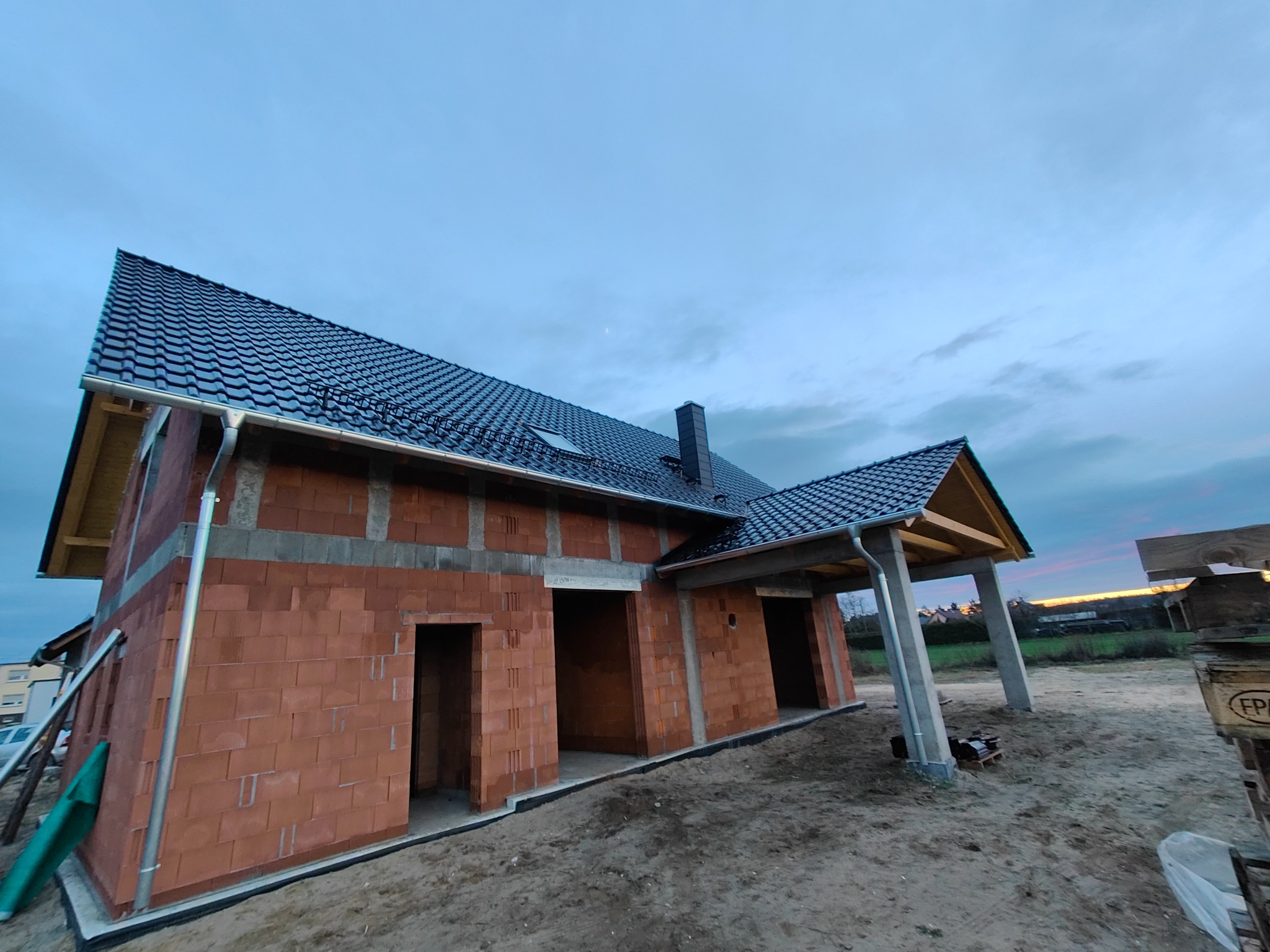 Budowa domu jednorodzinnego w stanie surowym otwartym, widoczny czarny dach z rynnami, ściany z czerwonej cegły, betonowe słupy podtrzymujące zadaszenie, tło wieczorne niebo.