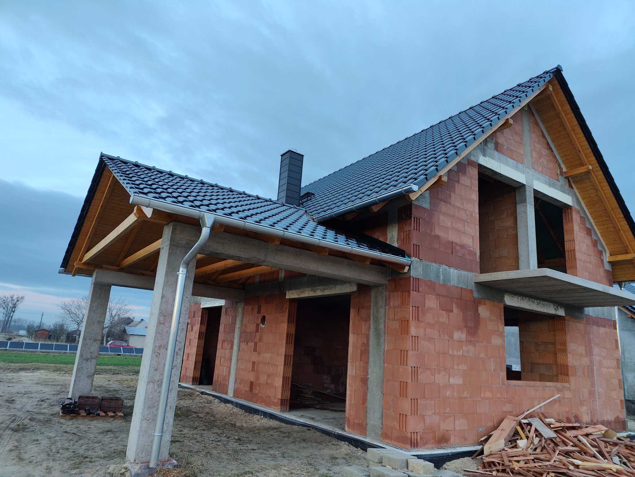 Surowy stan budynku jednorodzinnego z ciemną dachówką, widoczne elementy więźby, rynny i komin, ściany z czerwonej cegły, betonowe nadproża i słupy, teren budowy z resztkami drewna.