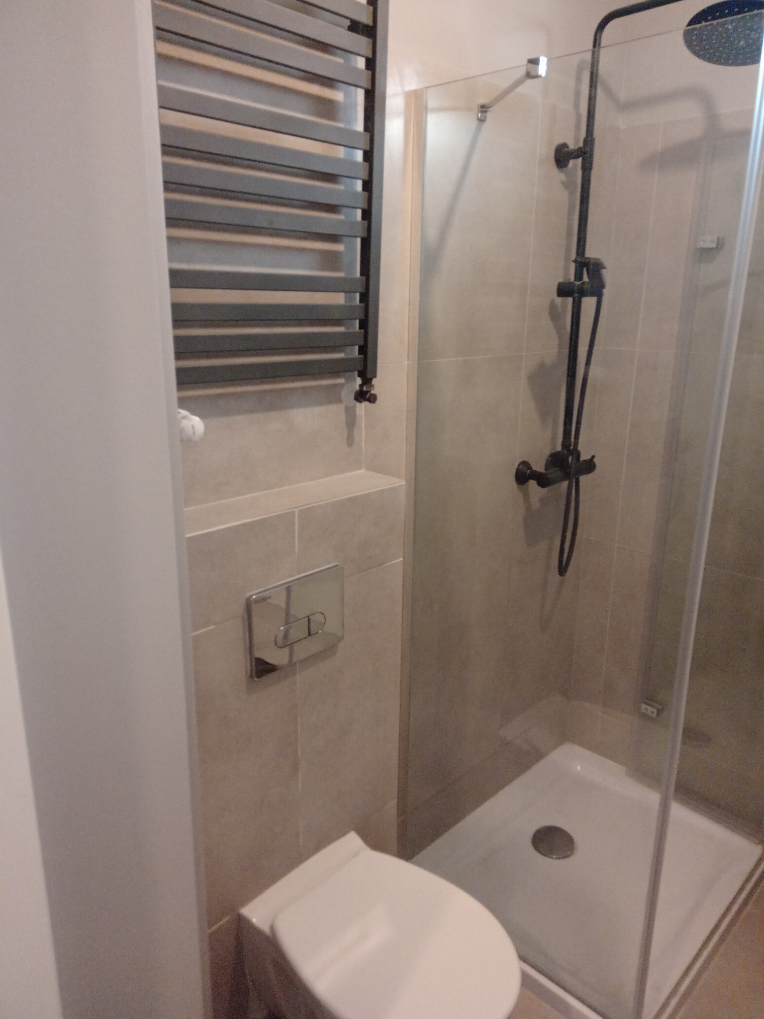 Nowoczesna łazienka z kabiną prysznicową typu walk-in, grzejnikiem drabinkowym w kolorze grafitowym, podwieszaną miską WC i beżowymi płytkami wielkoformatowymi.