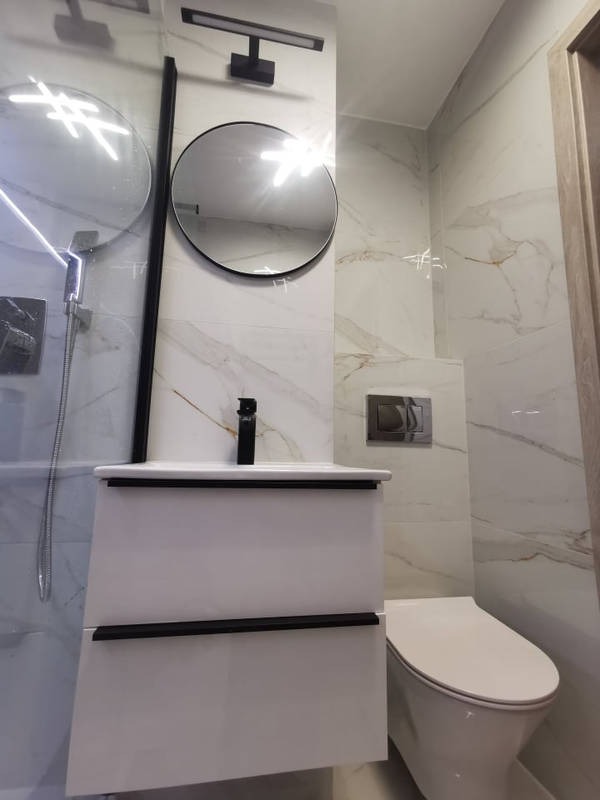 Nowoczesna łazienka z białymi marmurkowymi płytkami, okrągłym lustrem z czarną ramą, białą szafką pod umywalkę z czarnymi uchwytami i wiszącą toaletą.