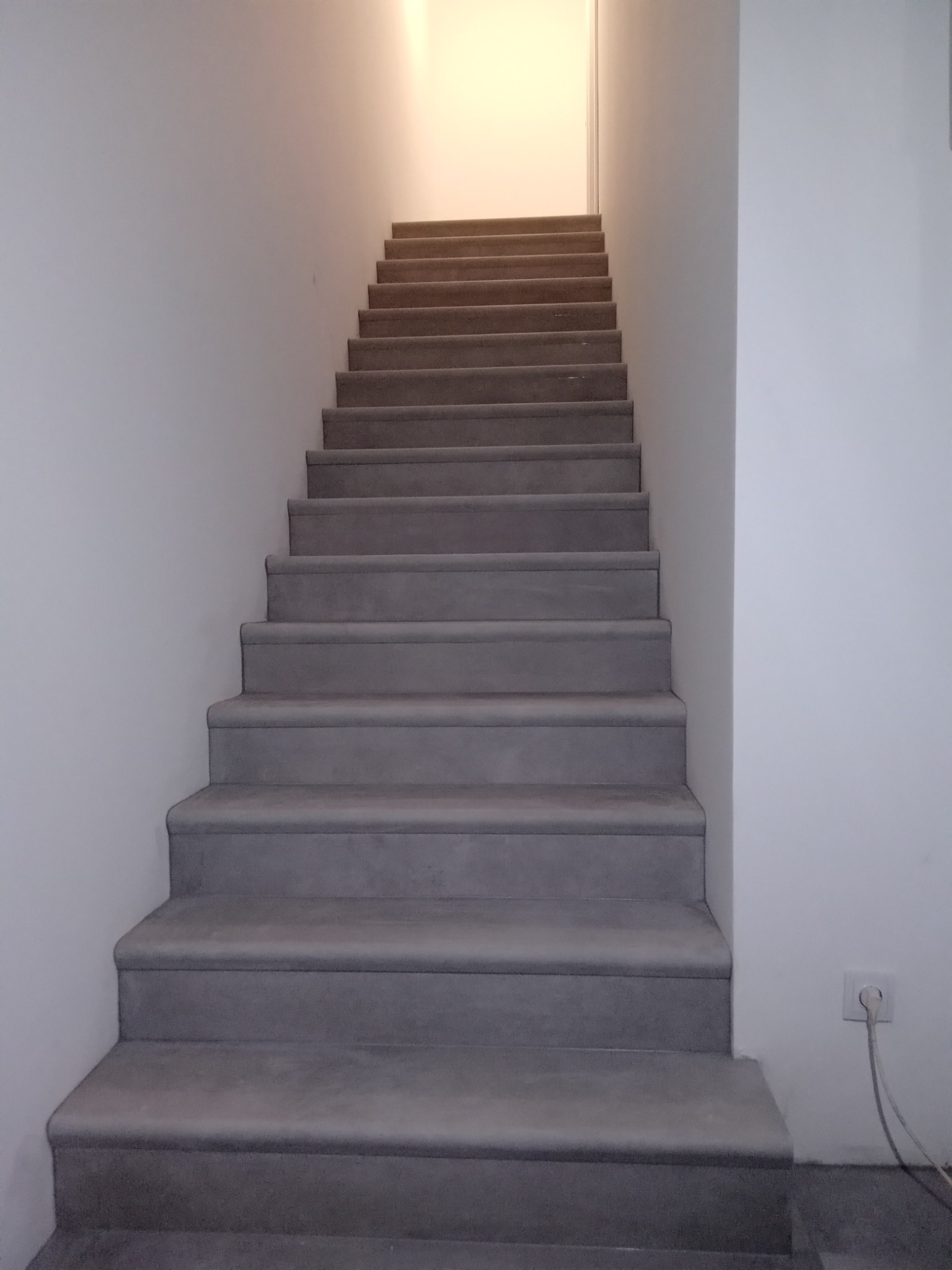 Schody betonowe prowadzące w górę, oświetlone od góry, z widocznym gniazdkiem elektrycznym na ścianie
