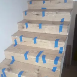 Schody wyłożone panelami drewnopodobnymi, zabezpieczone niebieską taśmą malarską na każdym stopniu, podczas prac wykończeniowych.