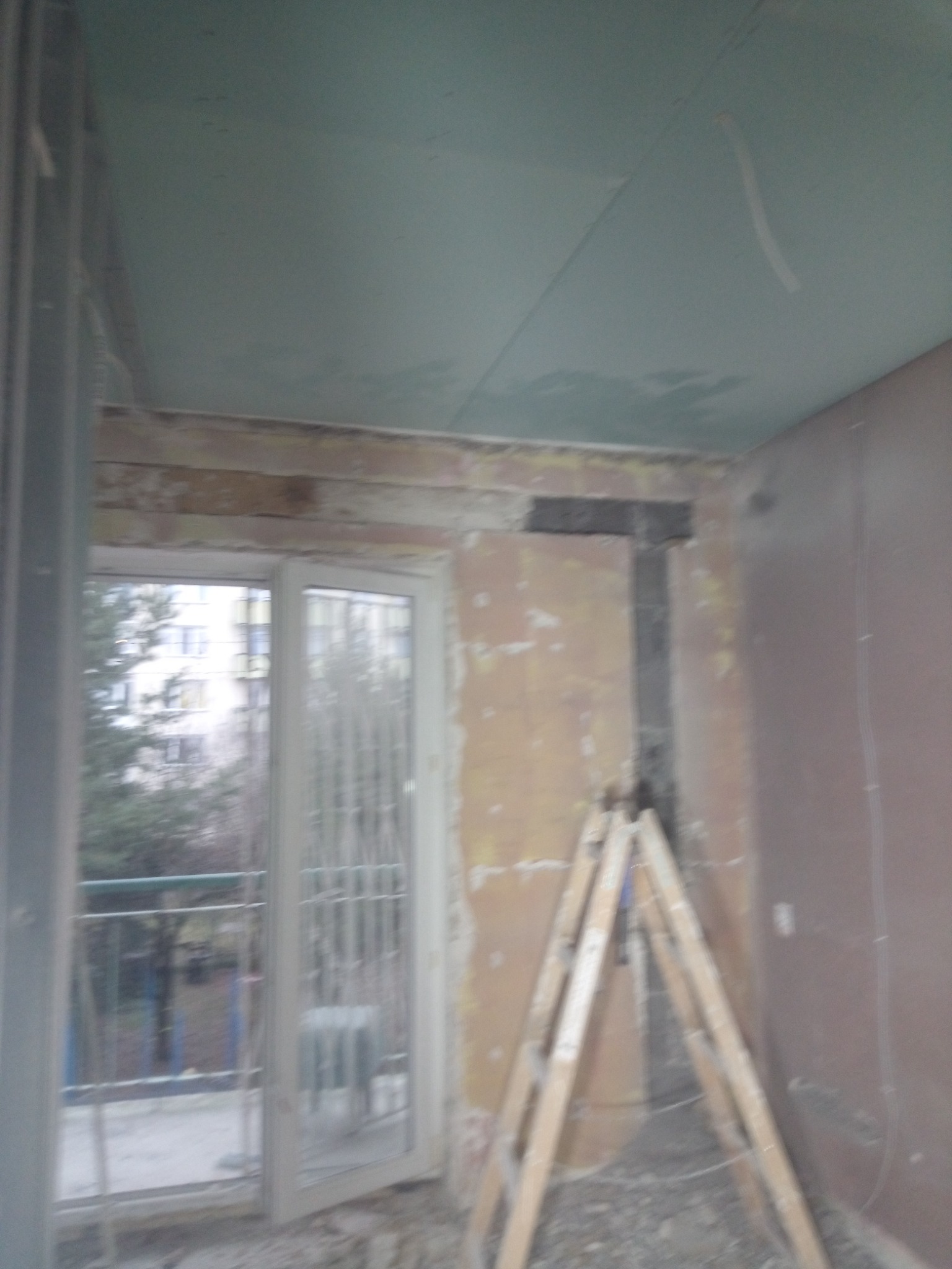 Pokój w trakcie prac przygotowawczych do malowania: widoczne ślady po starych tapetach, drabina oparta o ścianę, sufit z płyt kartonowo-gipsowych, okno balkonowe z widokiem na drzewa i budynek.