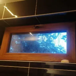 Wnęka w ścianie wyłożona czarnymi, błyszczącymi płytkami z jasną fugą, w której osadzone jest drewniane okno z widokiem na zaśnieżone drzewa.