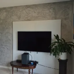 Ściana w salonie z efektem dekoracyjnej szarej faktury, widoczny telewizor, stolik z akcesoriami RTV oraz doniczkowa roślina.