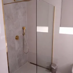 Szklana kabina prysznicowa z złotymi elementami wykończeniowymi, szare płytki imitujące kamień na ścianie, odpływ liniowy w podłodze, minimalistyczny design.