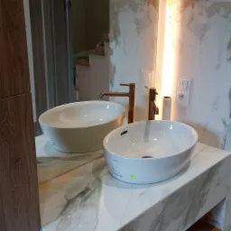 Dwie białe, owalne umywalki nablatowe z miedzianymi bateriami, osadzone na marmurowym blacie z półką, odbijające się w podświetlanym lustrze w łazience.