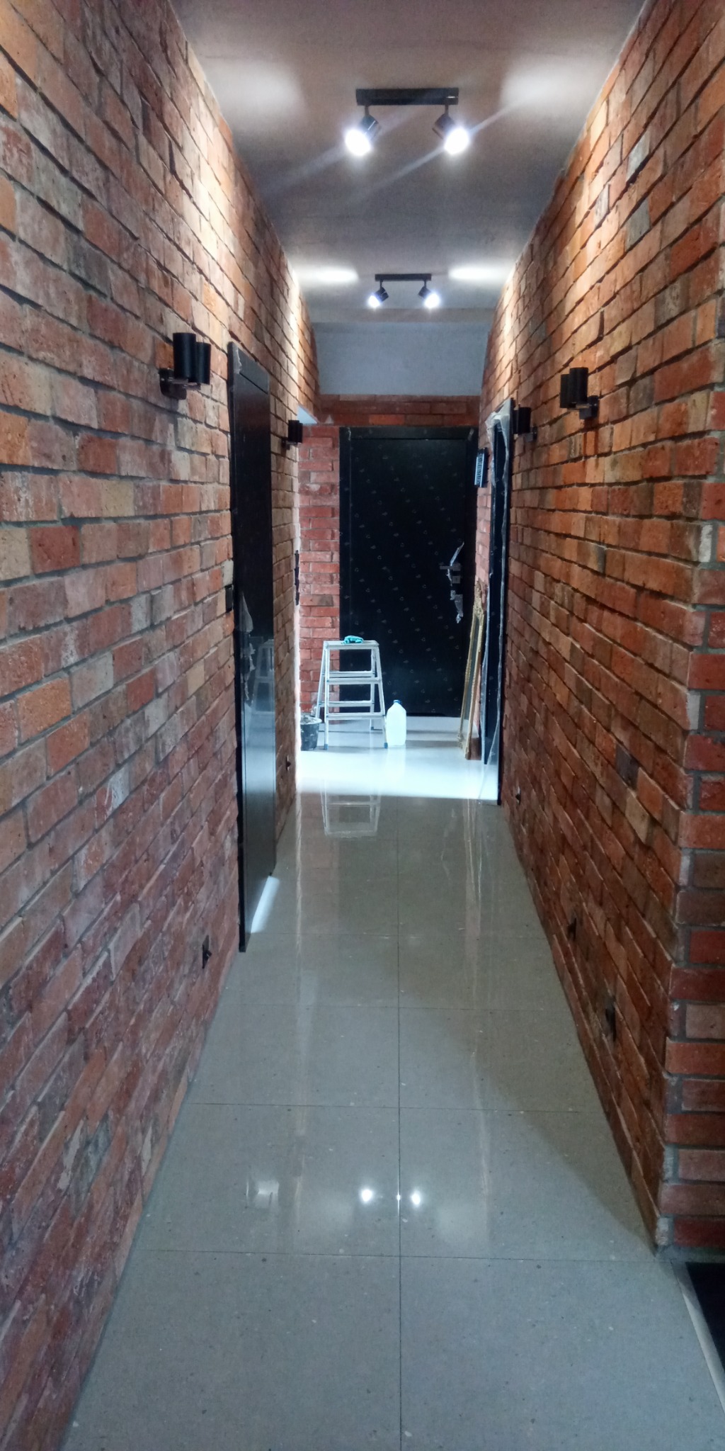 Wąski korytarz z ceglanymi ścianami, oświetleniem punktowym i błyszczącą podłogą z płytek, w tle drabina malarska i butelka wody.