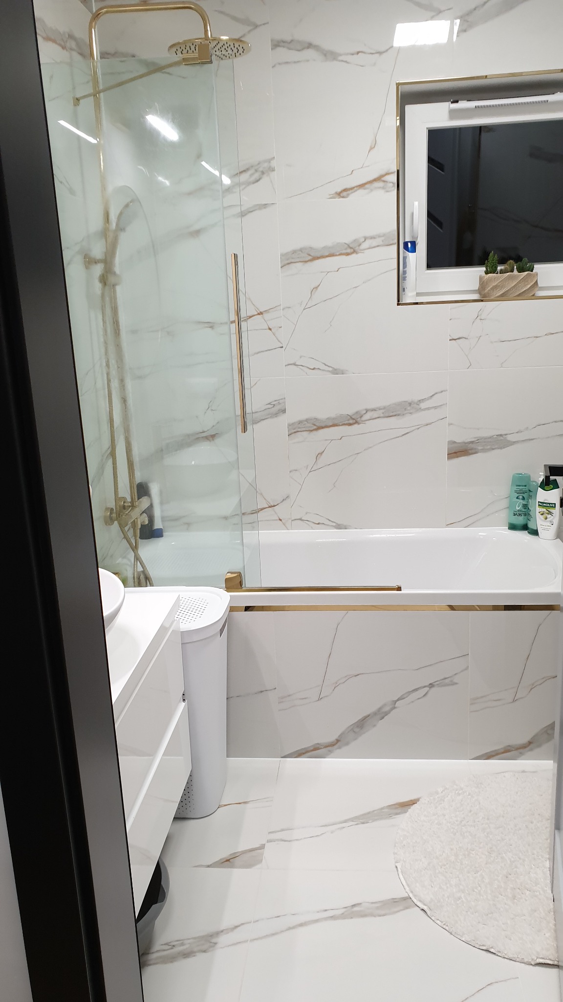 Nowoczesna łazienka z biało-złotymi płytkami imitującymi marmur, kabiną prysznicową ze złotą armaturą, białą wanną i okrągłym dywanikiem.