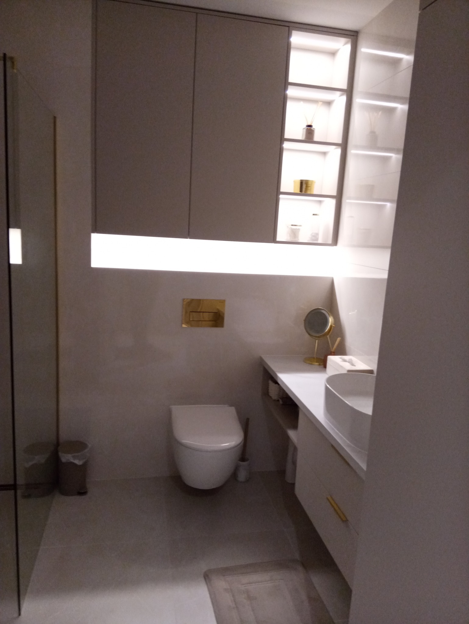 Nowoczesna łazienka w odcieniach szarości z podświetlanymi półkami i złotymi akcentami, widok na toaletę, umywalkę i szafki.