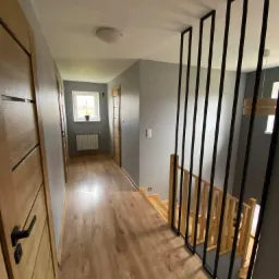 Wnętrze domu z drewnianą podłogą, szarymi ścianami, drewnianymi drzwiami z czarnymi klamkami oraz schodami z drewnianą balustradą i czarnymi metalowymi pionowymi elementami dekoracyjnymi.