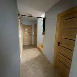 Wnętrze korytarza z szarą ścianą, drewnianymi drzwiami z poziomymi paskami i zamontowanym drążkiem do podciągania z kółkami gimnastycznymi.