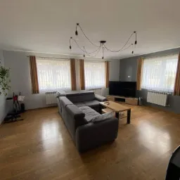 Jasne wnętrze salonu z drewnianą podłogą, szarą sofą narożną, telewizorem na komodzie i lampą sufitową z żarówkami na kablach.