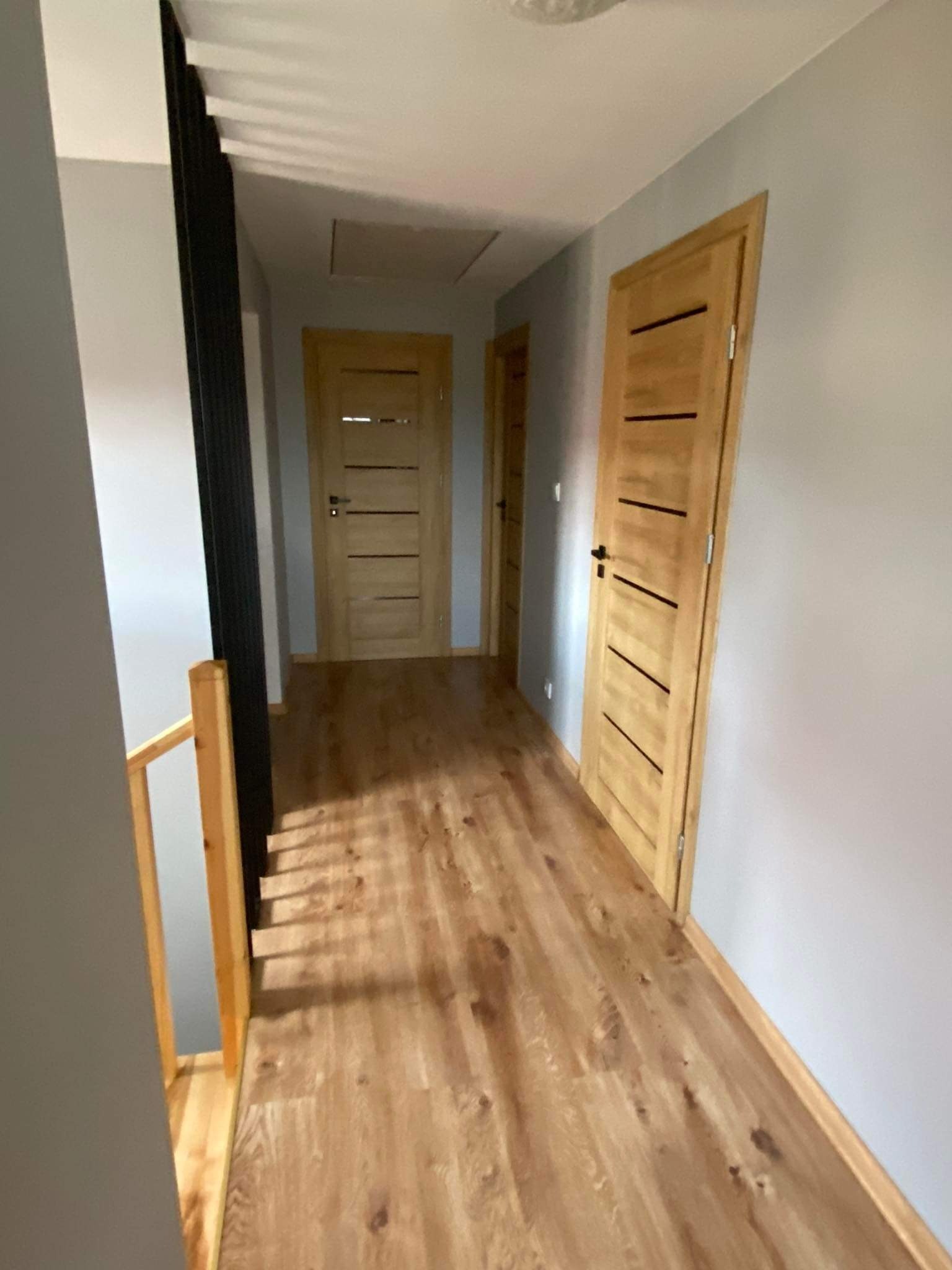 Korytarz z drewnianą podłogą, jasnymi drzwiami z ciemnymi akcentami i szarymi ścianami, widok z góry schodów z czarną balustradą.