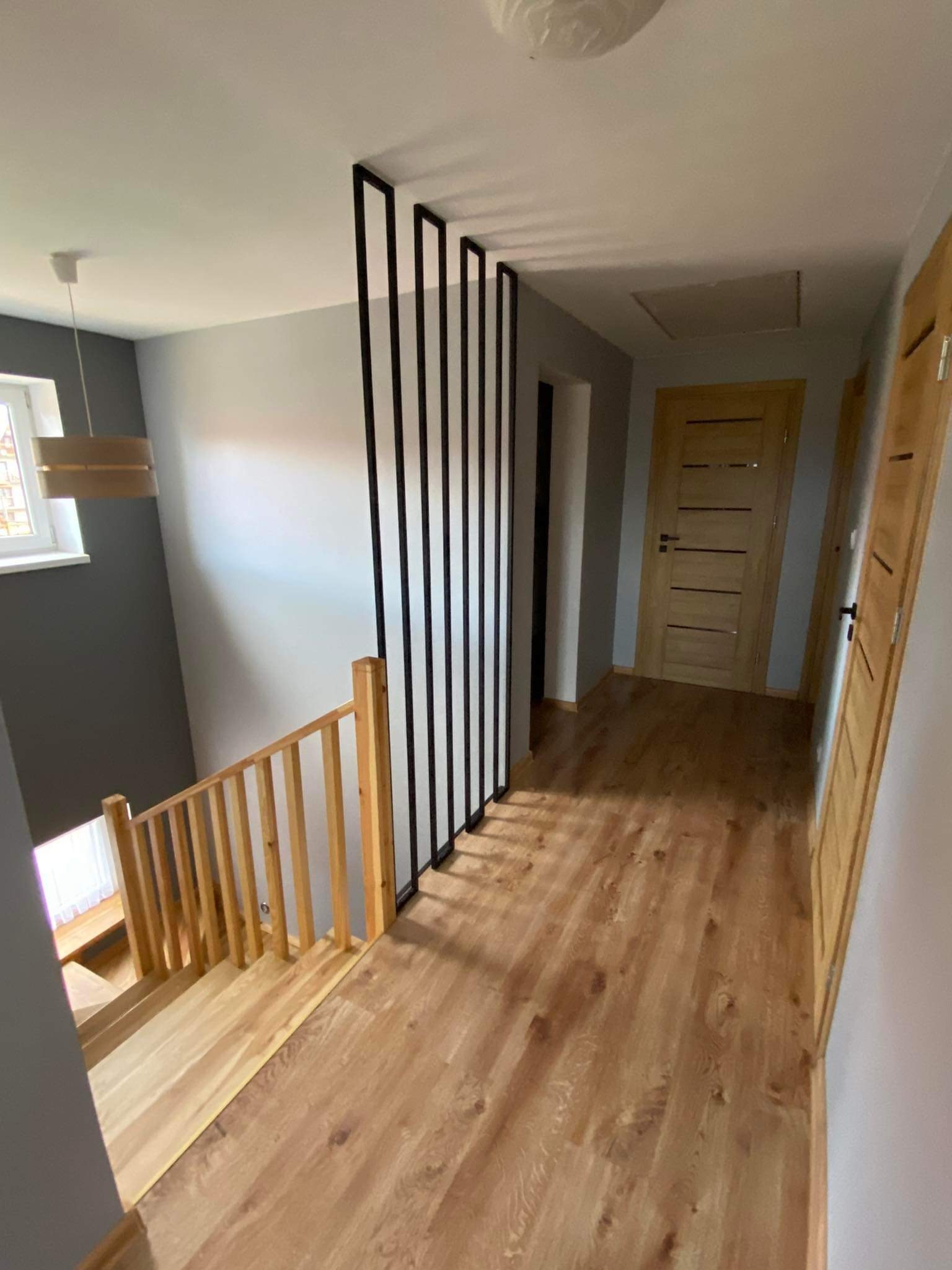 Korytarz z drewnianą podłogą i schodami, minimalistyczna czarna ścianka działowa z pionowych listew, widok z góry schodów, drzwi wewnętrzne z jasnego drewna.