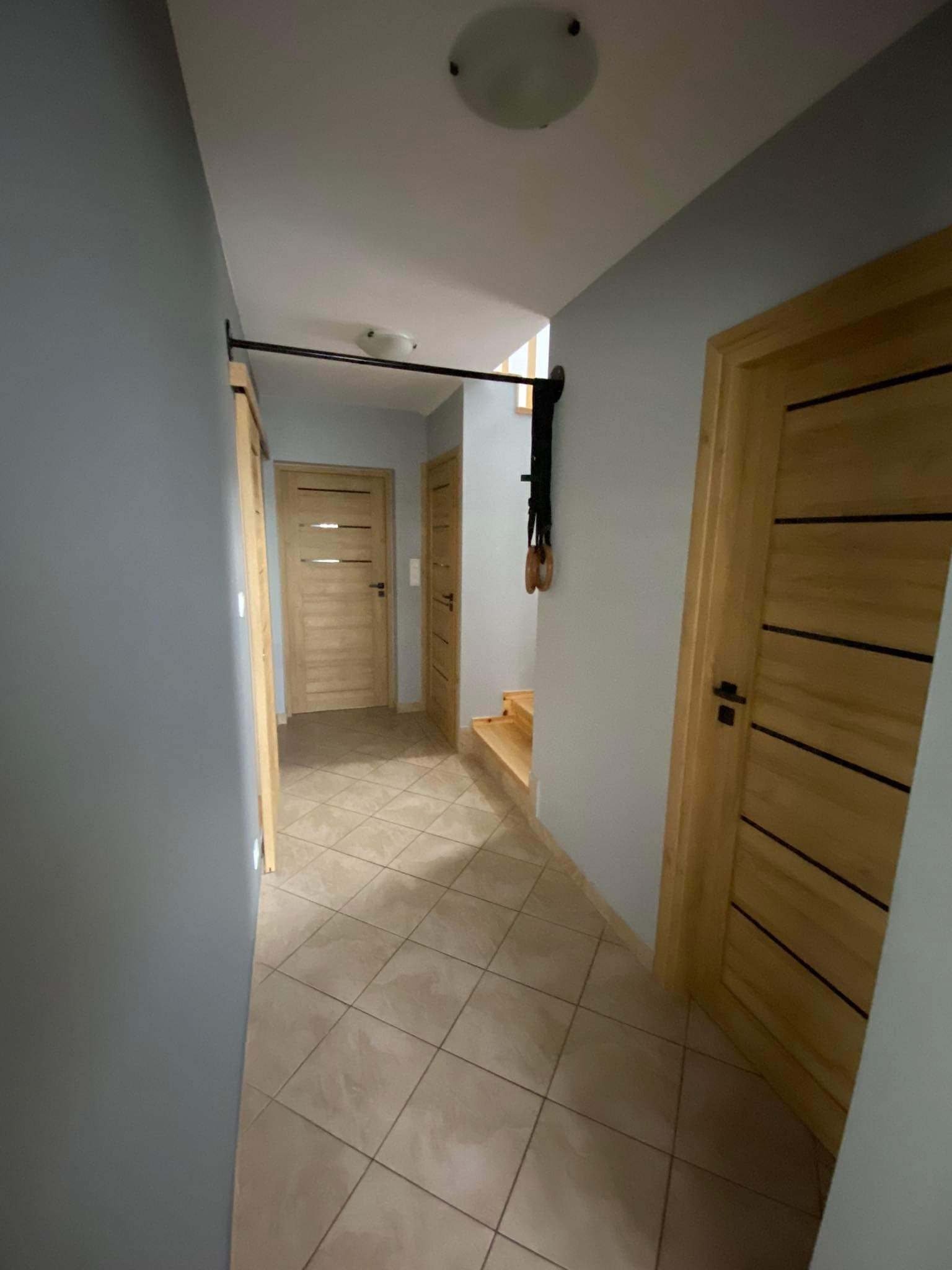 Wnętrze korytarza z szarą ścianą, drewnianymi drzwiami z poziomymi paskami i zamontowanym drążkiem do podciągania z kółkami gimnastycznymi.