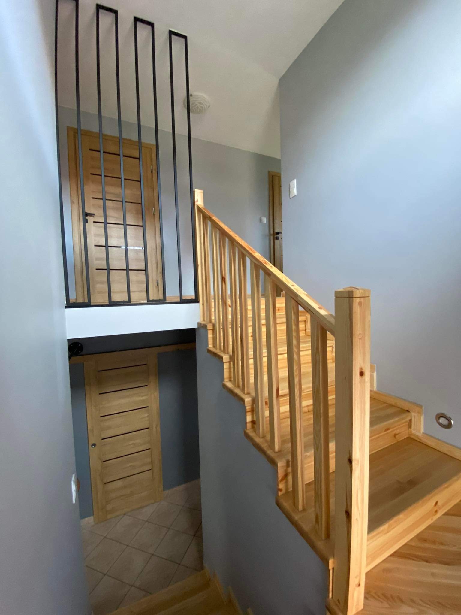 Jasne drewniane schody z balustradą w nowoczesnym wnętrzu z szarą ścianą i drzwiami z poziomymi panelami.