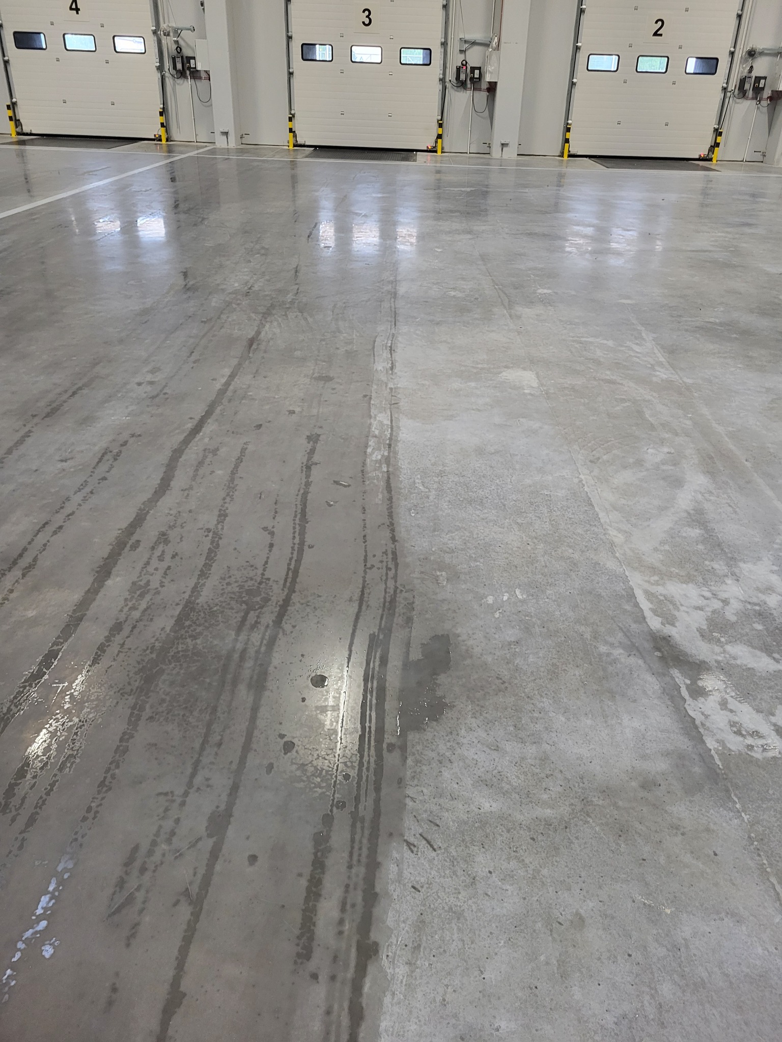 Polerowana, mokra posadzka betonowa w hali magazynowej z bramami w tle, ślady po myciu widoczne na powierzchni.