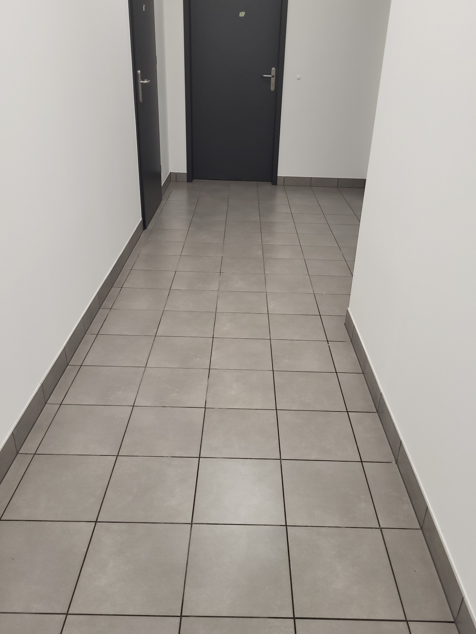Wnętrze korytarza biurowego z szarymi płytkami na podłodze, białymi ścianami i dwoma ciemnymi drzwiami.