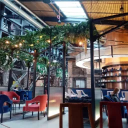 Wnętrze nowoczesnej restauracji z metalową konstrukcją stropu, dekoracjami roślinnymi, oświetleniem punktowym i barwnymi, welurowymi krzesłami w odcieniach czerwieni i granatu. W tle widoczny...