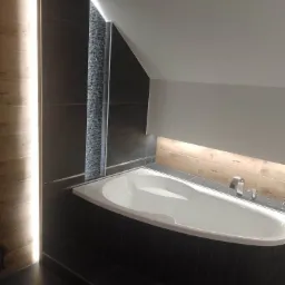 Nowoczesna łazienka na poddaszu z białą wanną obudowaną ciemnymi płytkami, oświetleniem LED wzdłuż ściany i pod półką, oraz dekoracyjnym panelem z kamienia naturalnego.