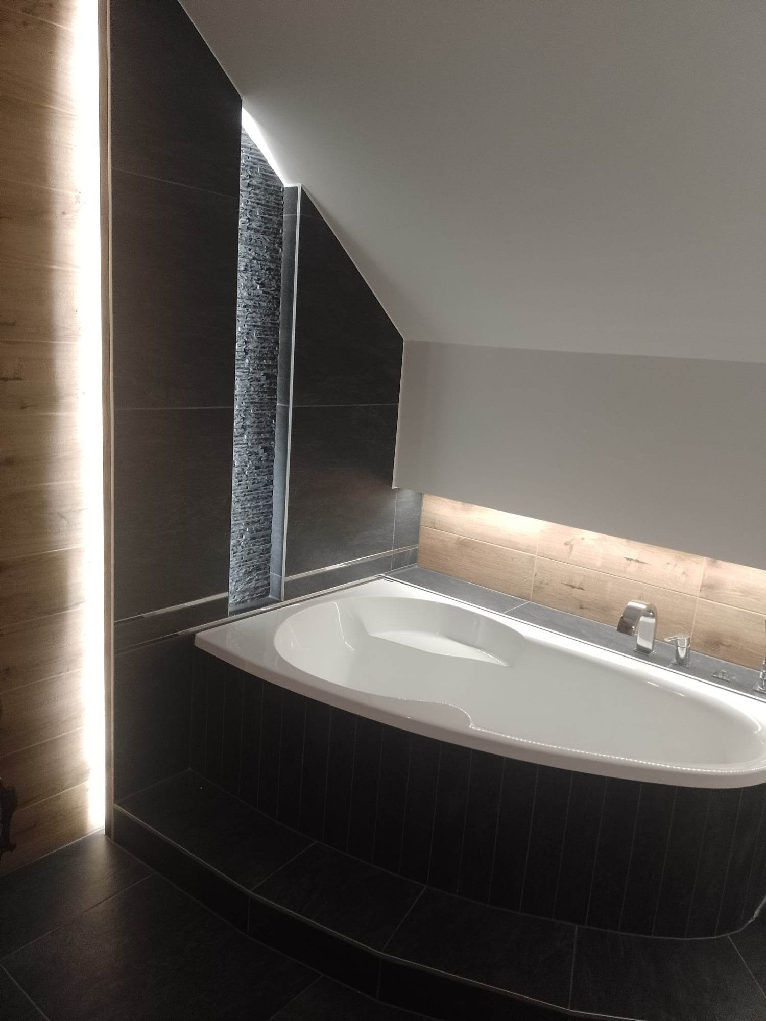 Nowoczesna łazienka na poddaszu z białą wanną obudowaną ciemnymi płytkami, oświetleniem LED wzdłuż ściany i pod półką, oraz dekoracyjnym panelem z kamienia naturalnego.