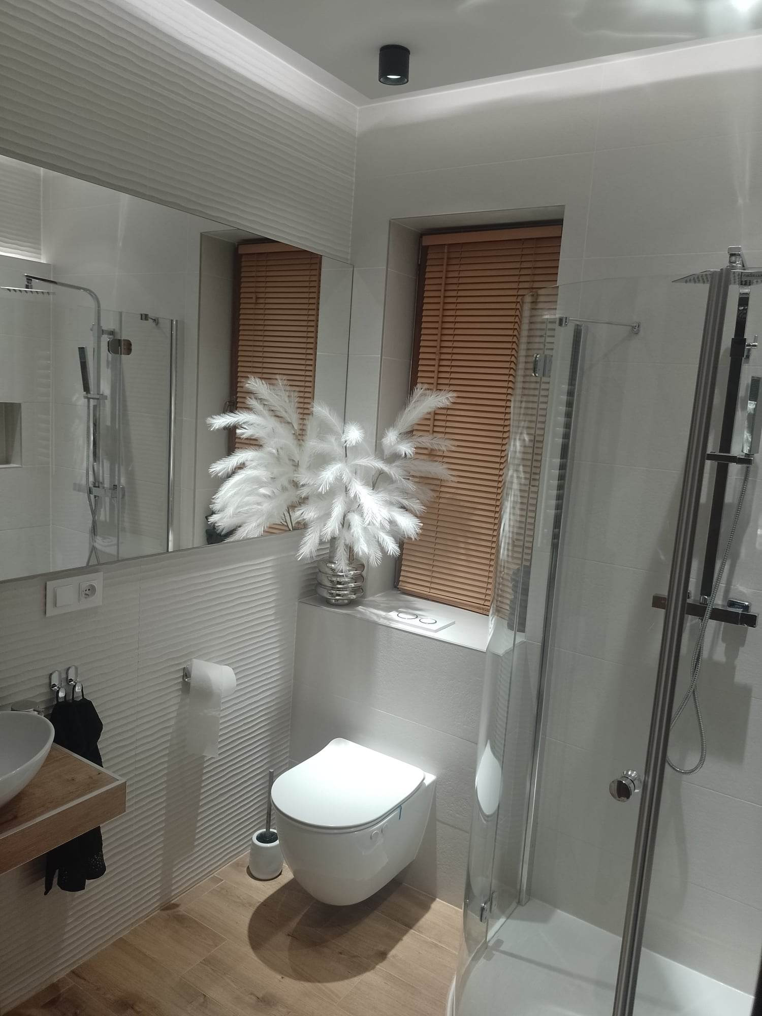 Nowoczesna łazienka z drewnianymi akcentami: podłoga w odcieniach drewna, umywalka na drewnianym blacie, żaluzje w oknie, biała armatura i dekoracyjne trawy pampasowe w srebrnym wazonie.