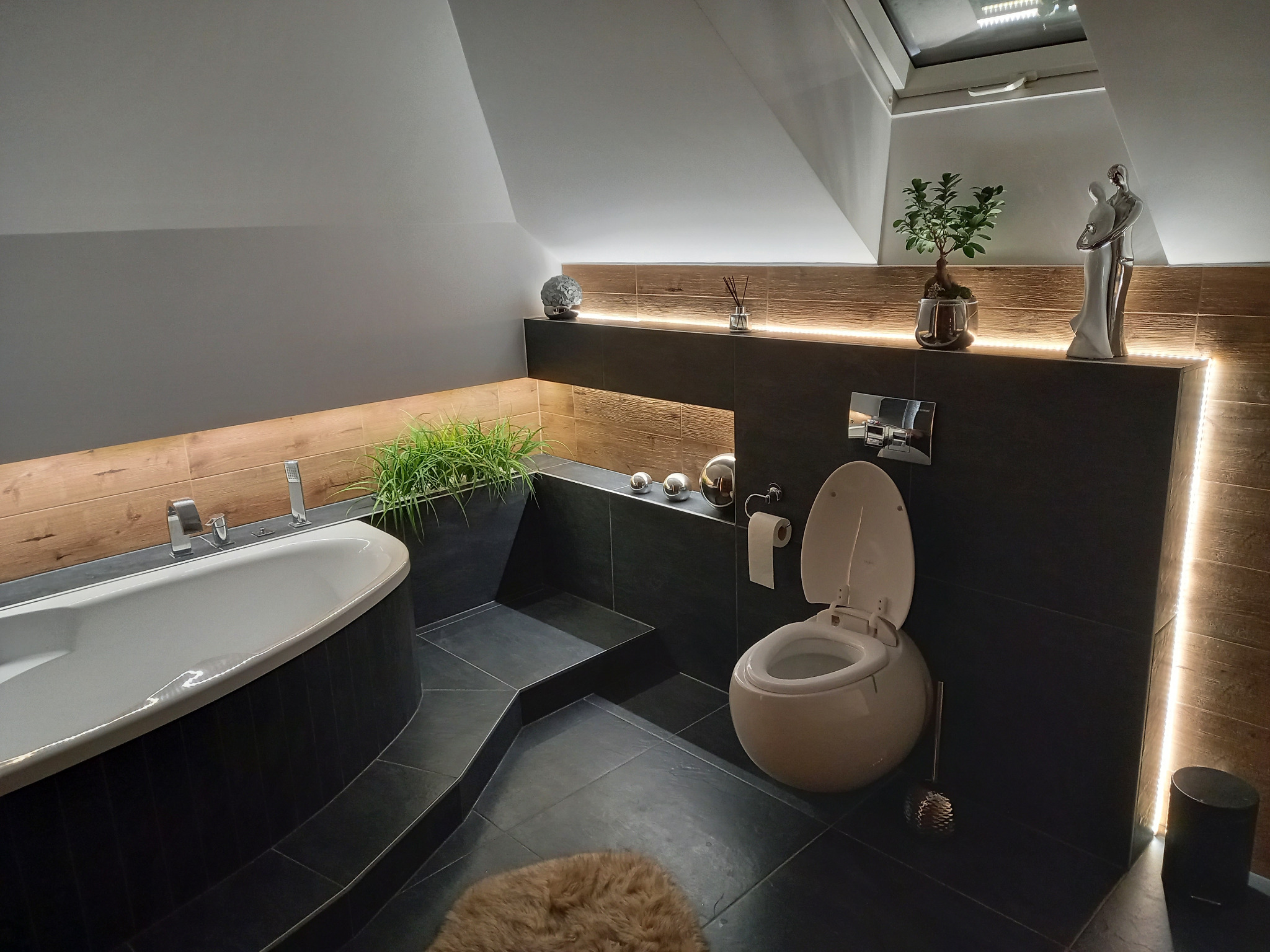 Stylowa łazienka na poddaszu z wanną, toaletą z otwartą deską, czarnymi płytkami i drewnianymi akcentami podświetlonymi LED, uzupełniona roślinami i dekoracjami.