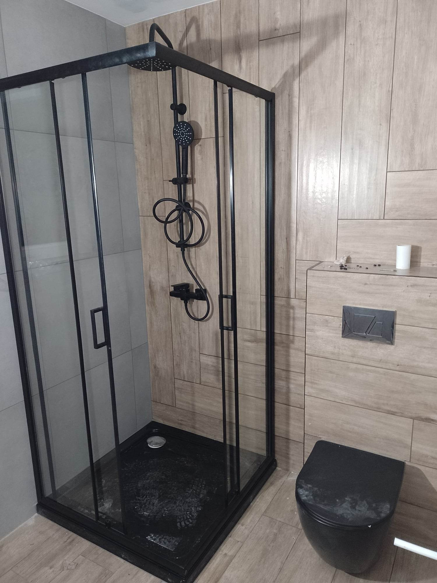 Kabina prysznicowa z czarną armaturą i brodzikiem, widoczna toaleta w kolorze czarnym, oraz płytki imitujące drewno na ścianach, zrealizowane w Tarnowie.