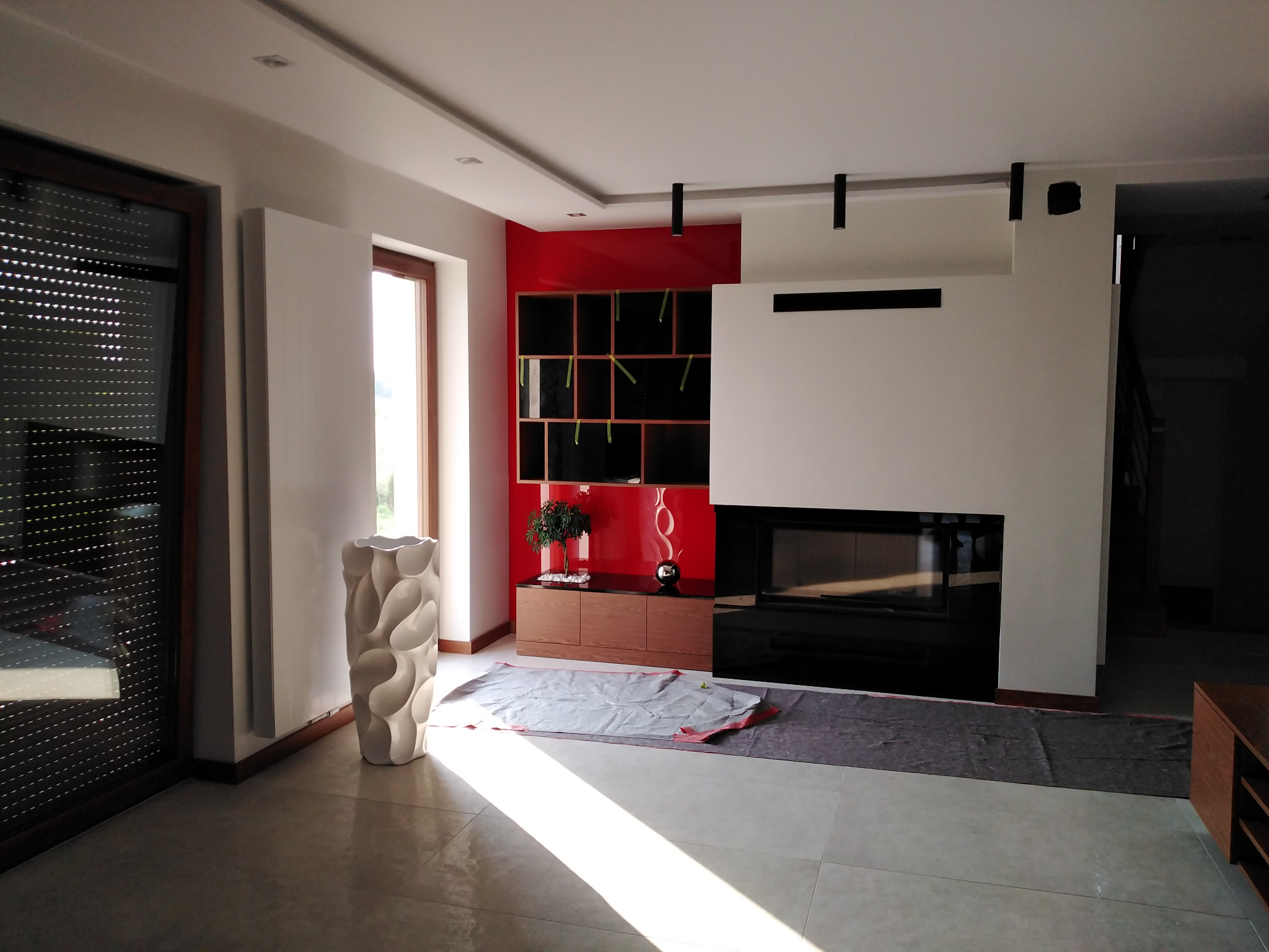 Nowoczesny salon z czerwonym akcentem na ścianie, minimalistycznym kominkiem i designerską wazą. Podłoga zabezpieczona folią malarską podczas prac.