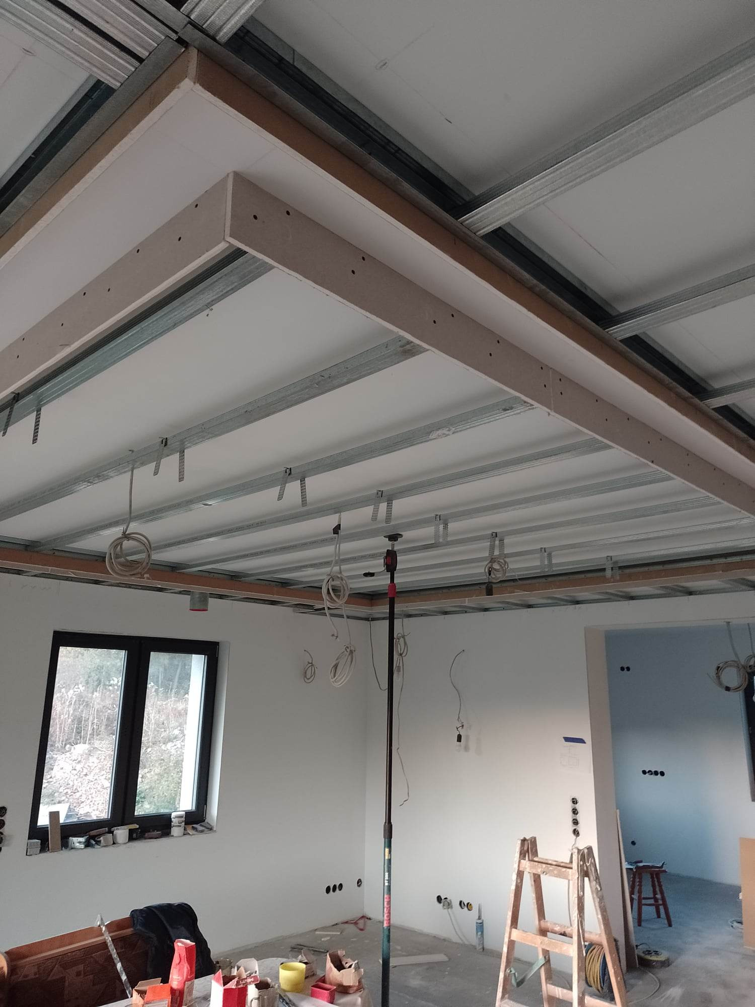 Sufit podwieszany w trakcie montażu, widoczne metalowe profile, płyty gipsowo-kartonowe oraz drewniana konstrukcja obramowania, okablowanie wystające z sufitu, drabina w tle.