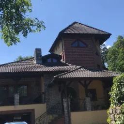 Dwupiętrowy dom z ciemną dachówką, ceglaną wieżą i elementami z kamienia, widoczny z perspektywy ulicy w słoneczny dzień.