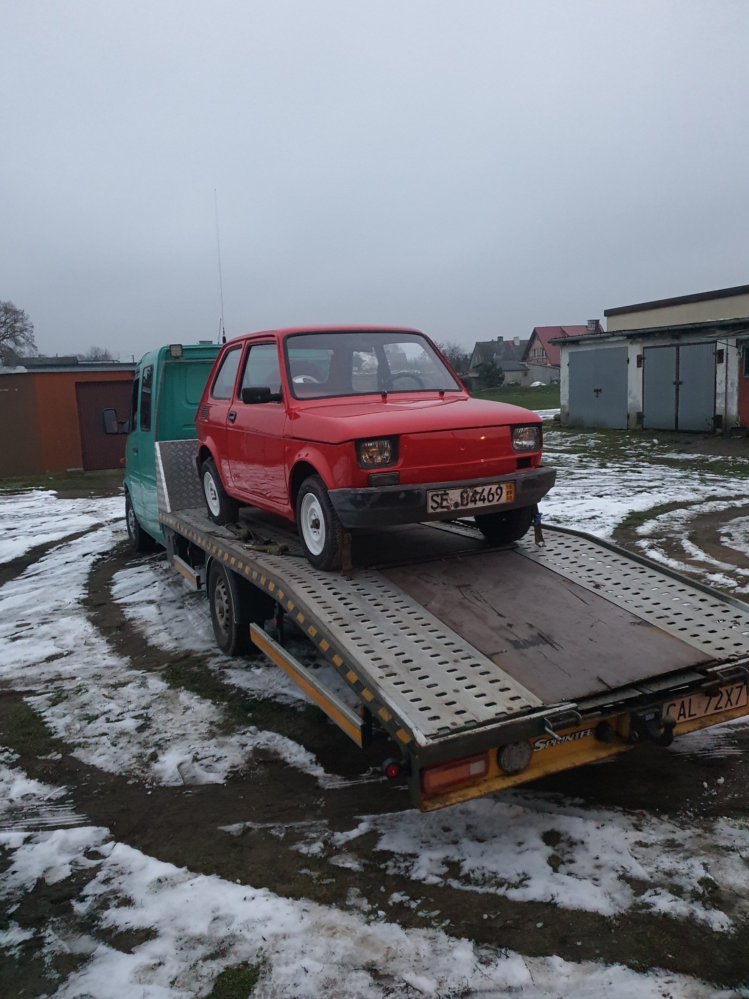 Czerwony Fiat 126p na platformie lawety, widok z przodu, na tle zimowego krajobrazu z zabudowaniami.