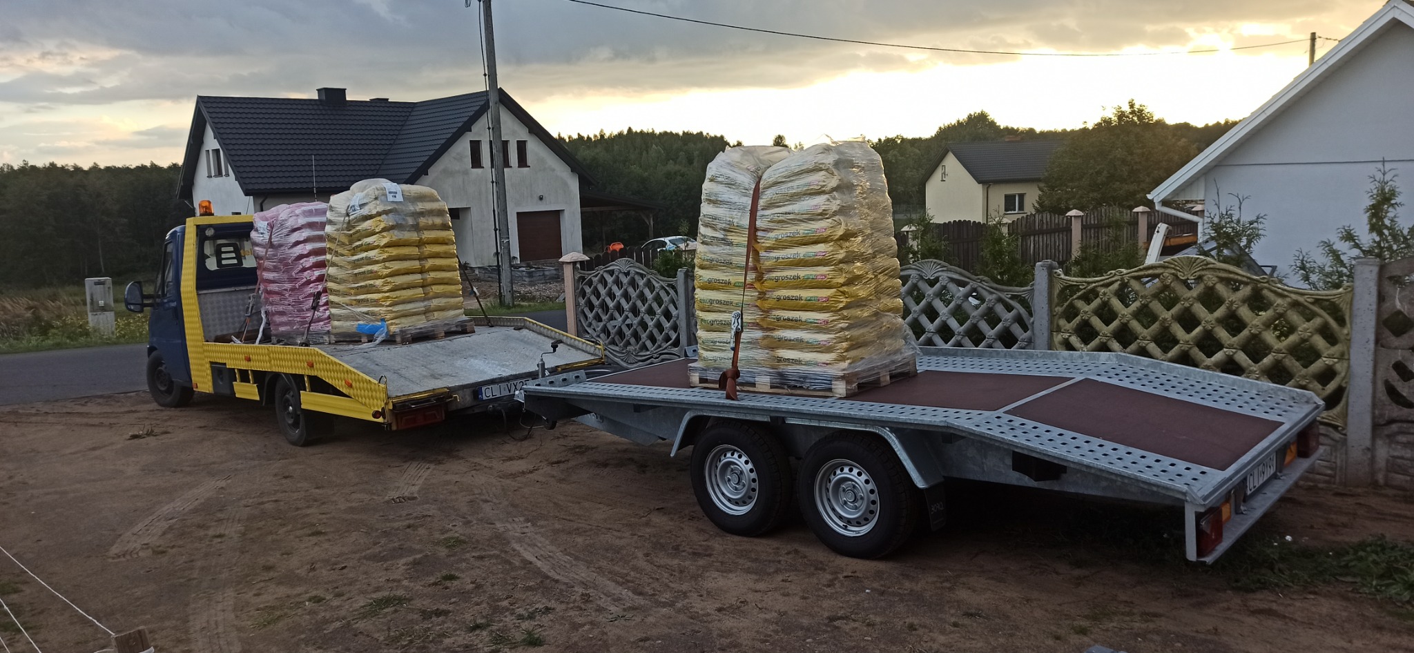 Żółto-niebieski bus z przyczepą lawetą przewozi palety z workami na tle zabudowań mieszkalnych.