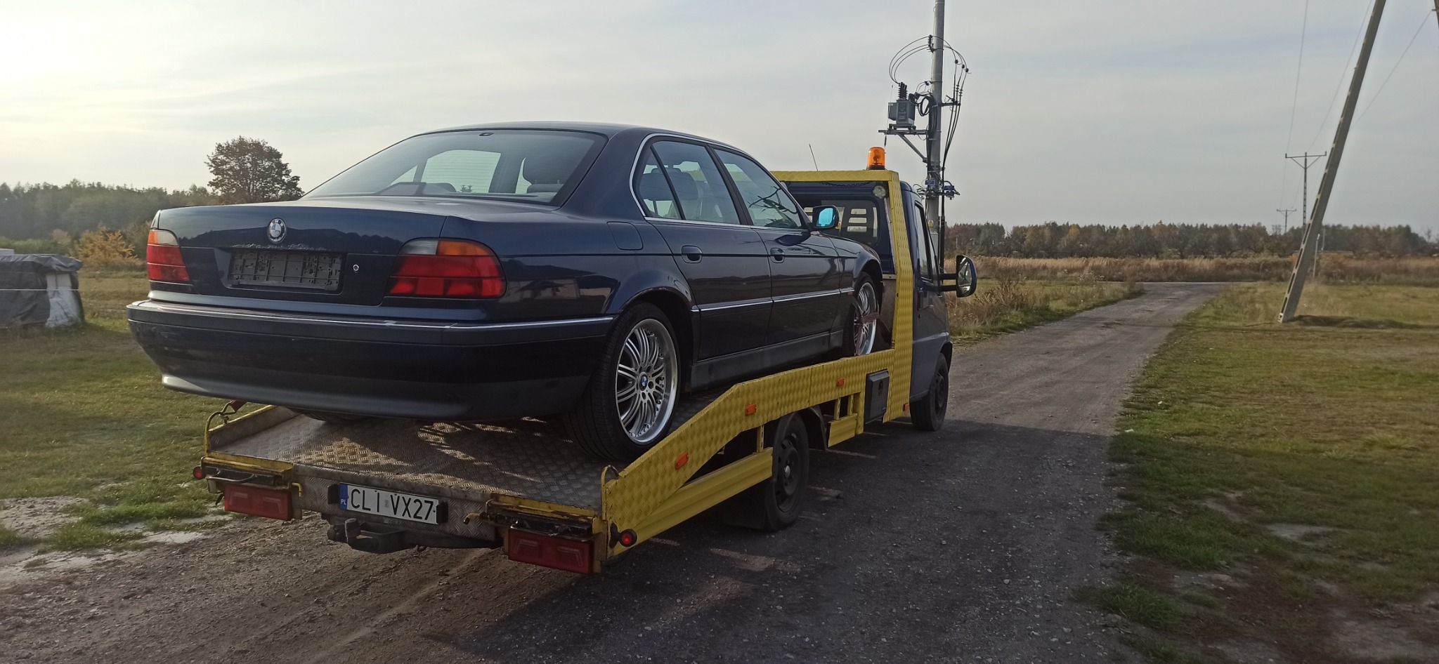 Granatowa limuzyna BMW serii 7 na żółtej lawecie, widok z tyłu, tablica rejestracyjna CLI VX27, polne otoczenie.