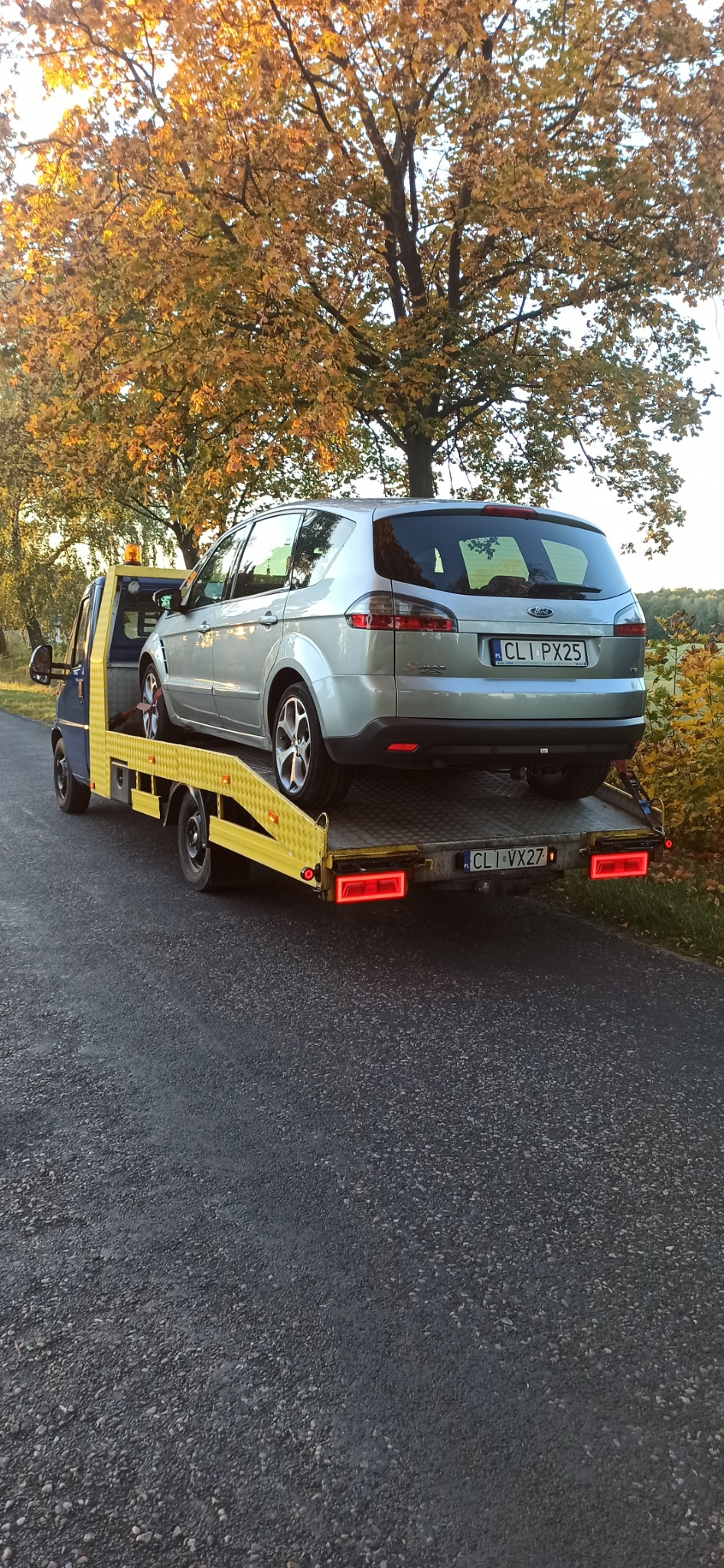 Srebrny samochód osobowy marki Ford S-Max z polskimi tablicami rejestracyjnymi przewożony na żółtej lawecie na tle jesiennego krajobrazu z drzewami o żółtych liściach.