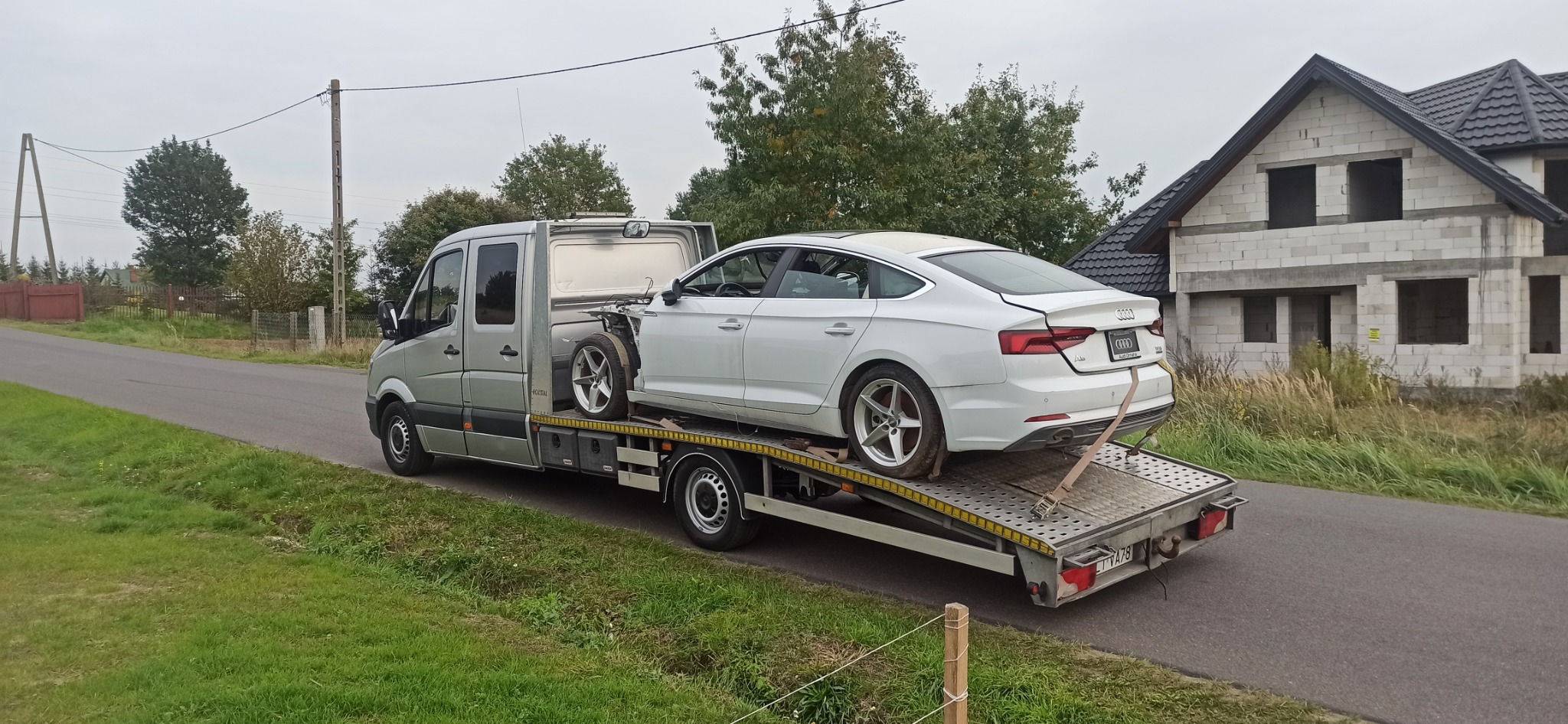 Laweta przewozi uszkodzone białe Audi A5 na tle niedokończonego domu jednorodzinnego.