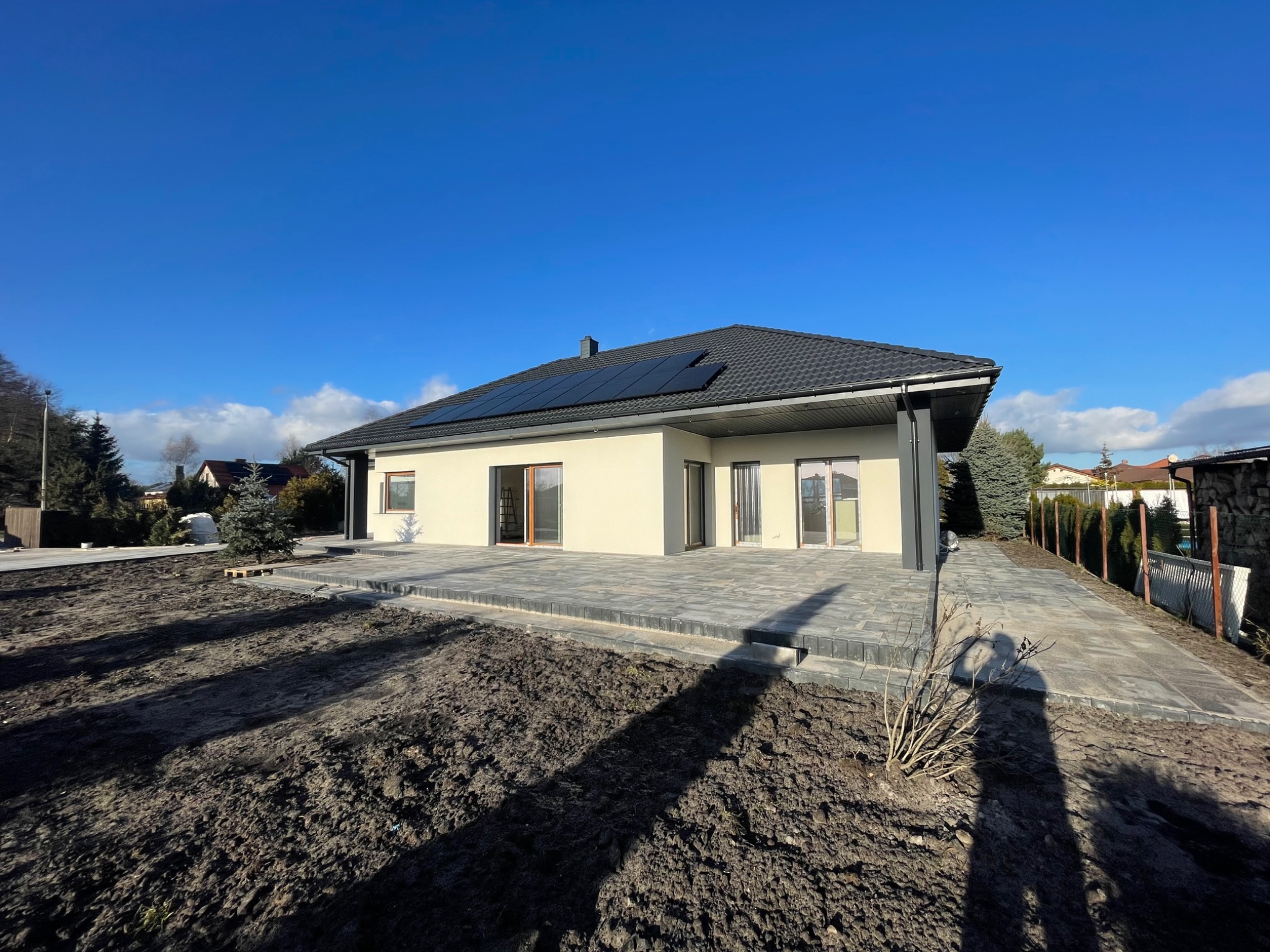 Nowoczesny dom jednorodzinny z panelami słonecznymi na dachu, wykończony jasnym tynkiem, z tarasem wyłożonym szarą kostką brukową, na tle błękitnego nieba.