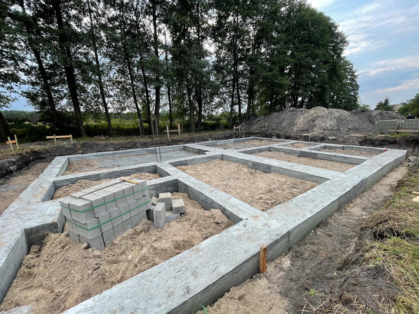 Wylane fundamenty domu jednorodzinnego z widocznym zbrojeniem i stertą bloczków betonowych przygotowanych do dalszych prac murarskich, w tle drzewa i usypana ziemia.