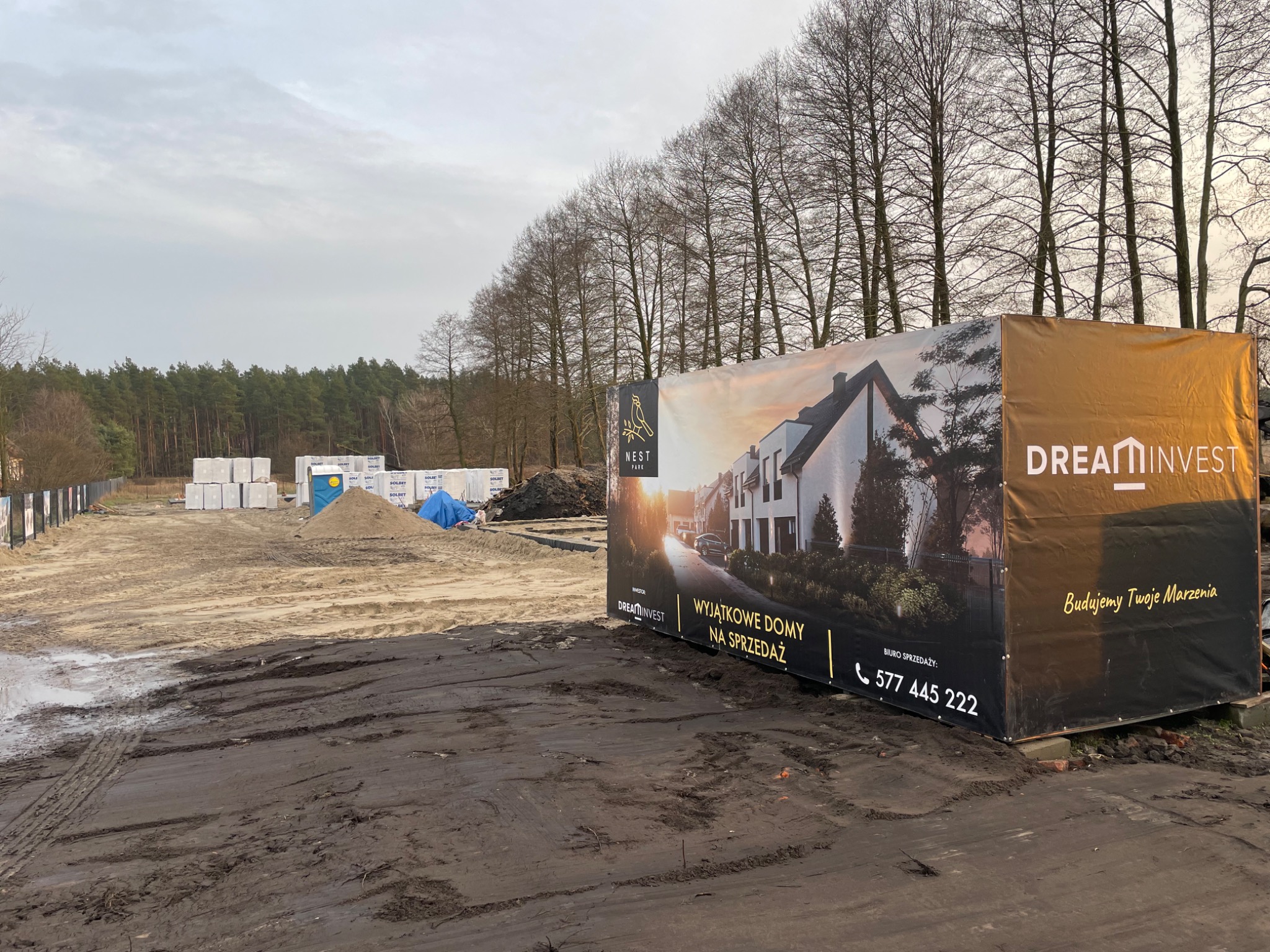 Plac budowy z widocznymi materiałami budowlanymi, ziemią i banerem reklamowym z wizualizacją nowoczesnego domu oraz logo firm budowlanych Dream Invest i Nest Park.